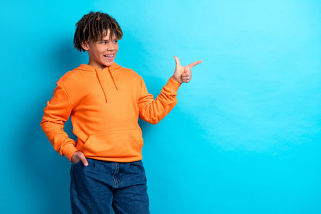 Cheerful young boy in an orange hoodie making a pointing gesture against a vibrant blue backgroundの写真素材
