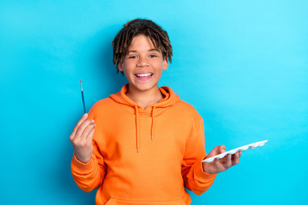 Happy young boy holding a paintbrush and palette in orange hoodie on a blue background expressing creativityの写真素材