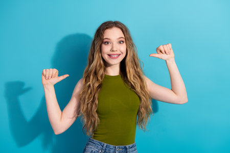 Cheerful young woman in a green top pointing to herself on a vibrant blue background, expressing confidence and happinessの写真素材
