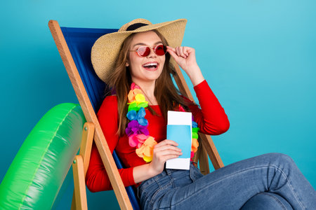 Cheerful young woman in a summer outfit sitting on a wooden chair with travel accessories and...