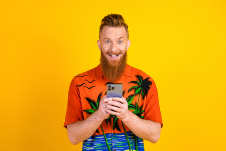 Cheerful young man with red hair in colorful tropical shirt using smartphone against vibrant yellow...
