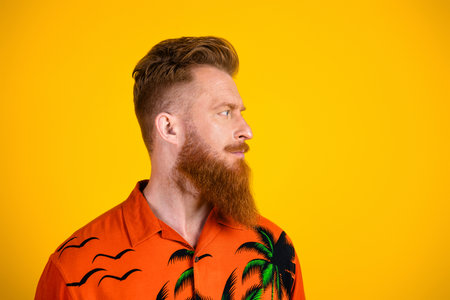 Portrait of a stylish young man with a tropical print shirt and red beard against a vibrant yellow...