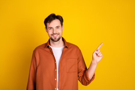 Young man in casual orange shirt points to copy space with a cheerful gesture concept on yellow backgroundの写真素材