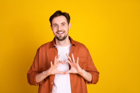 Handsome happy man in orange shirt forming a heart shape with his hands against a vibrant yellow...