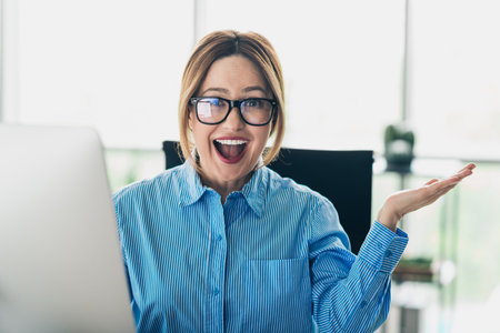 Young businesswoman in a bright office setting looking cheerful while engaging with work on her computerの写真素材