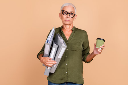Confident mature businesswoman carrying work files, holding coffee on beige background, portraying...