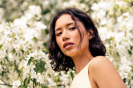 Beautiful young woman surrounded by blooming flowers in a sunny outdoor setting, showcasing gentle charm and natural beautyの写真素材