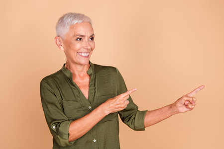 Smiling senior woman with short grey hair pointing joyfully, showcasing positivity and charm in an elegant casual khaki shirtの写真素材