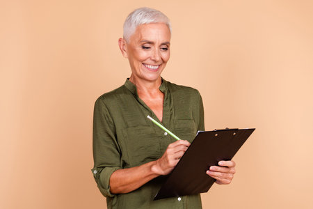 Smiling mature woman with short grey hair holding a clipboard and pencil in her hand against a beige backgroundの写真素材