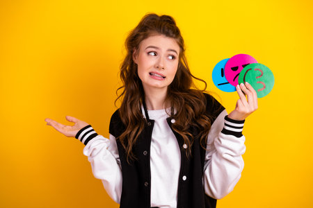 Cute woman holding emotional expression cards against a vibrant yellow background showing puzzled gestureの写真素材