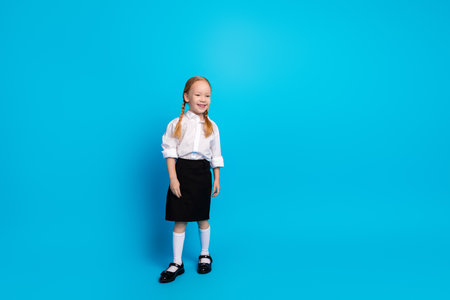 Charming red-haired schoolgirl posing against a bright blue background in a classic school uniform, smiling with joyの写真素材