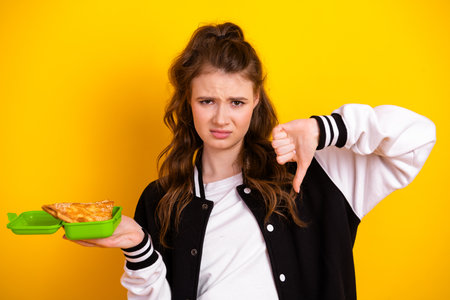 Teen girl in casual outfit expressing displeasure at food in a green container with a thumbs-down gesture and yellow backdropの写真素材