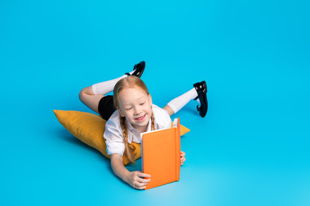 Young schoolgirl with braids lying on a pillow while reading an orange book, on a bright blue...