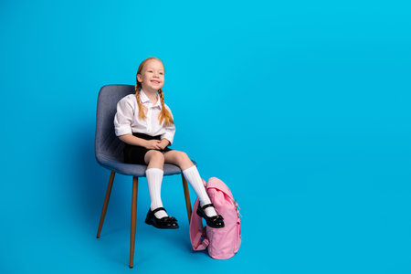 Smiling young girl with red braids sitting in uniform with backpack on blue background embodying school spiritの写真素材