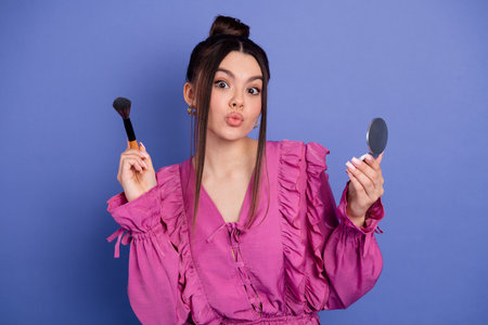 Beautiful young woman in a pink ruffled dress holding a makeup brush and mirror against a purple backgroundの写真素材