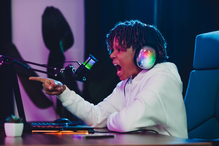 Excited young gamer wearing headphones streaming online in a vibrant home studio setup with neon lightingの写真素材