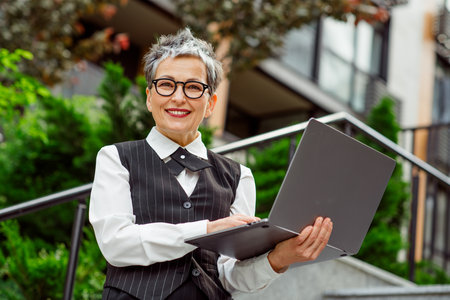 Elegant Professional Woman Using Laptop Outdoors in Urban Setting Showing Confidence and Style with a Bright Smileの写真素材