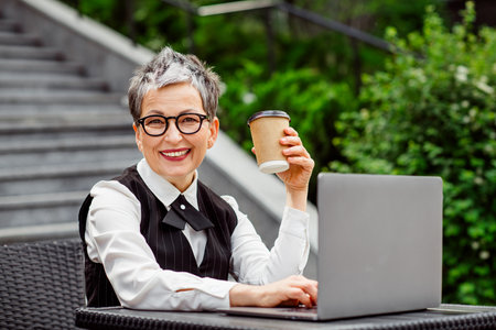 Smiling businesswoman working outdoors and enjoying coffee on a sunny day, showcasing elegance, confidence, and charmの写真素材