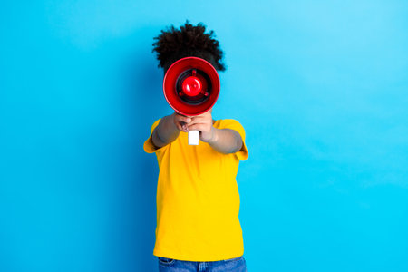 Young girl with afro hairstyle using a megaphone, wearing a yellow shirt, on a vibrant blue background, promoting ideasの写真素材