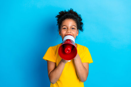 Cheerful young girl holding a red megaphone on a bright blue background, wearing a yellow t-shirt,...