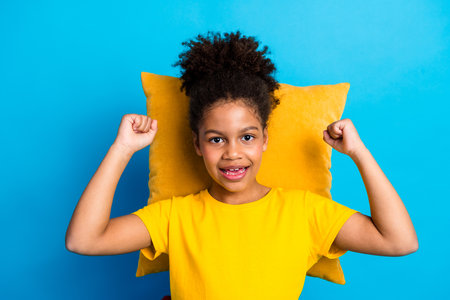 Cheerful young girl posing against blue background with yellow pillow and vibrant outfit showcasing positive energyの写真素材