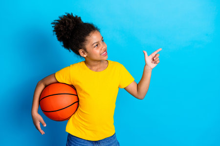 Cheerful young girl holding a basketball in a casual outfit, pointing proudly with a bright expression, on a blue backgroundの写真素材