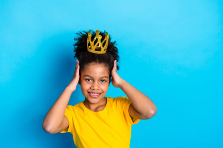Cheerful young girl wearing a golden crown accessory over a blue backdrop, expressing joy and confidenceの写真素材