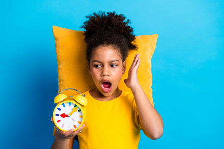 Young girl startled while holding a yellow clock and sitting against a pillow on a vibrant blue backgroundの写真素材