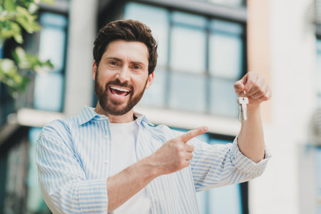Young bearded man smiling with keys outdoors next to modern building, representing themes of real estate success and happinessの写真素材