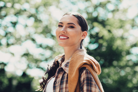 Portrait of a smiling young woman in casual attire enjoying a sunny day outdoors against a backdrop of vibrant foliageの写真素材