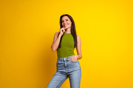 Charming woman in a green top and jeans posing against a vibrant yellow background, expressing confidence and happinessの写真素材