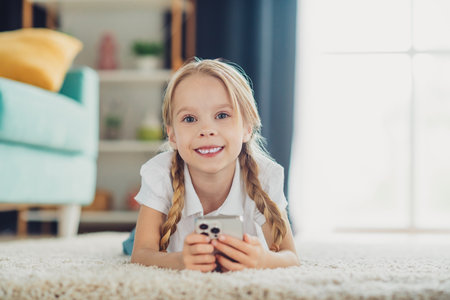 Adorable young girl with braids smiling while holding a smartphone, enjoying leisure time in a modern cozy living roomの写真素材