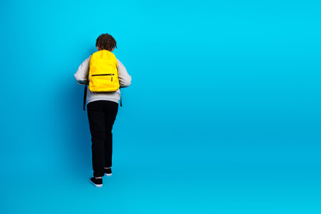 Student wearing a yellow backpack standing against a blue background showcasing education and casual styleの写真素材
