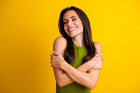 Smiling young brunette female in green top embracing herself against yellow backdrop, showcasing positivity and styleの写真素材