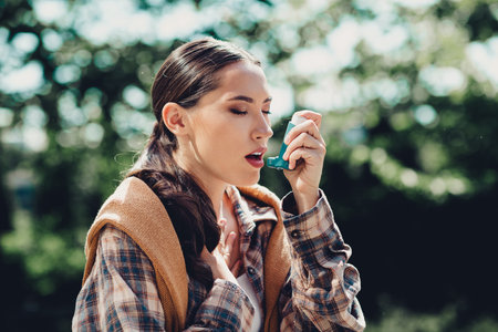 Young woman outdoors using an inhaler during an asthma episode on a bright sunny day in a green parkの写真素材