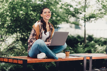 Young woman enjoying outdoor productivity with a laptop seated on a park bench surrounded by vibrant greeneryの写真素材