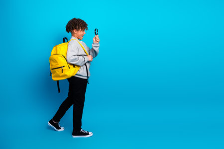Smiling schoolboy with bright yellow backpack playing with a magnifying glass against a blue background, cheerful studentの写真素材