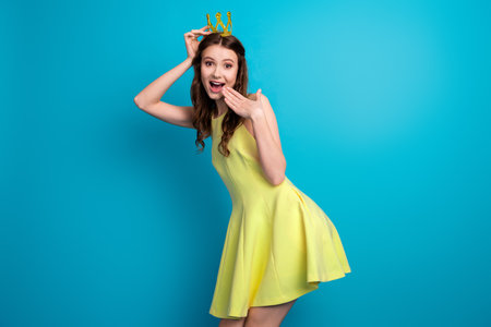 Joyful young brunette wearing a yellow dress and golden crown against a bright blue background expressing happiness and charmの写真素材