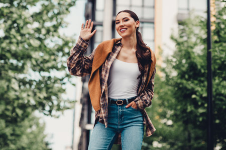 Attractive young woman waving and smiling outdoors during casual urban walk on a sunny day in a modern city streetの写真素材