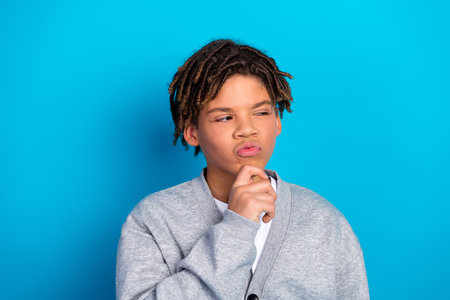 Charming schoolboy pondering with hand gesture against a blue background wearing a casual cardigan...