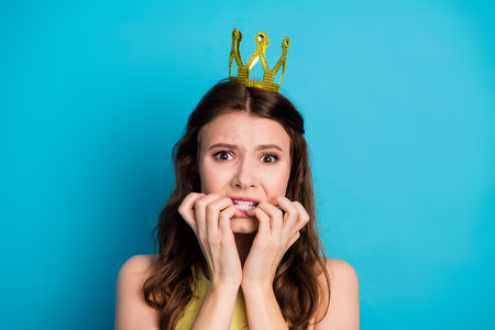 Charming young woman in a golden crown expressing nervousness against a vivid blue backdropの写真素材