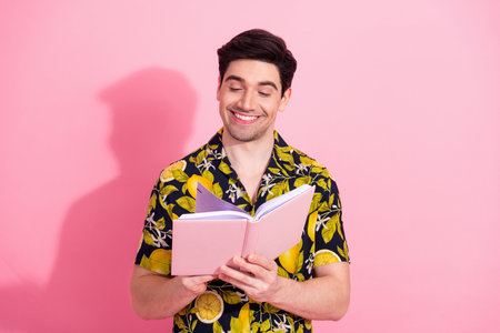 Young fashionably dressed man in tropical shirt reading a notebook with smile against a vibrant pink...
