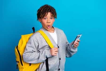 Excited schoolboy holding smartphone with surprise expression showcasing youth and education on vibrant blue backgroundの写真素材