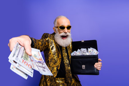 Charismatic senior man in vibrant attire holding cash and gesturing in sleek pose against vivid purple backgroundの写真素材