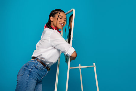 Young woman in casual outfit posing joyfully against vibrant blue background holding a modern mirrorの写真素材