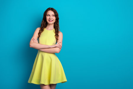 Young woman in a vibrant yellow dress posing confidently with arms crossed against a cheerful blue backgroundの写真素材
