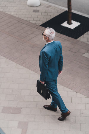 Professional businessman in a formal suit walking outdoors in an urban cityscape holding a briefcase.の写真素材