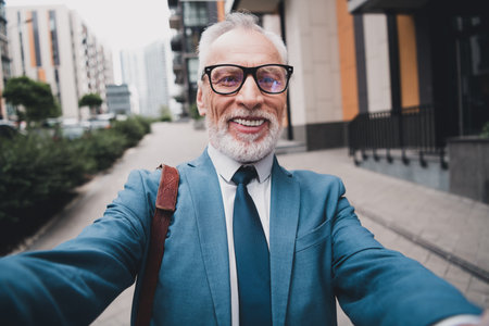 Successful senior businessman taking a cheerful selfie outdoors in an urban modern city street while wearing a formal suitの写真素材