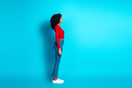 Side profile view of young woman in red top with curly hair on vibrant blue background showcasing casual fashionの写真素材