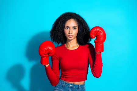 Young woman in vibrant red attire wearing boxing gloves poses confidently against a bright turquoise...